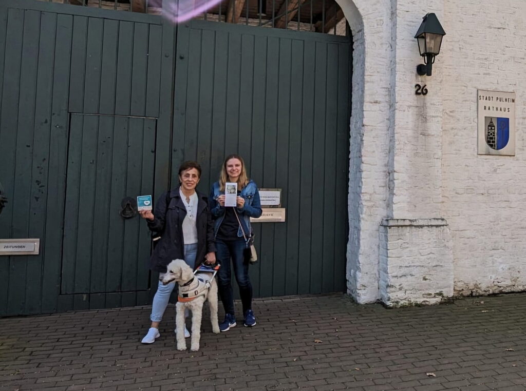 Marisa Sommer mit ihrem Blindenführhund Rainer und Sandra Fiedler (Initiative "Pulheim für Hunde") verteilen die Aufkleber "Assistenzhund Willkommen" in Pulheim, um ein Zeichen für Inklusion zu setzen.